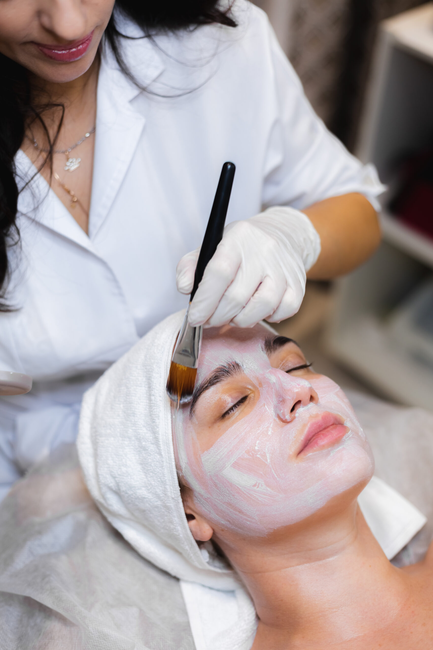 Beautician with a brush applies a white moisturizing mask to the face of a young girl client in a spa beauty salon